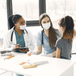 Duas médicas de máscara acompanham uma criança durante um atendimento numa sala hospitalar.