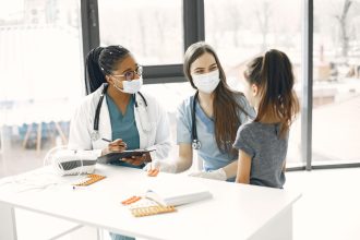 Duas médicas de máscara acompanham uma criança durante um atendimento numa sala hospitalar.
