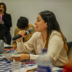 Mulher participa numa discussão de grupo, segurando um microfone junto a uma mesa.