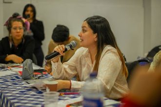 Mulher participa numa discussão de grupo, segurando um microfone junto a uma mesa.
