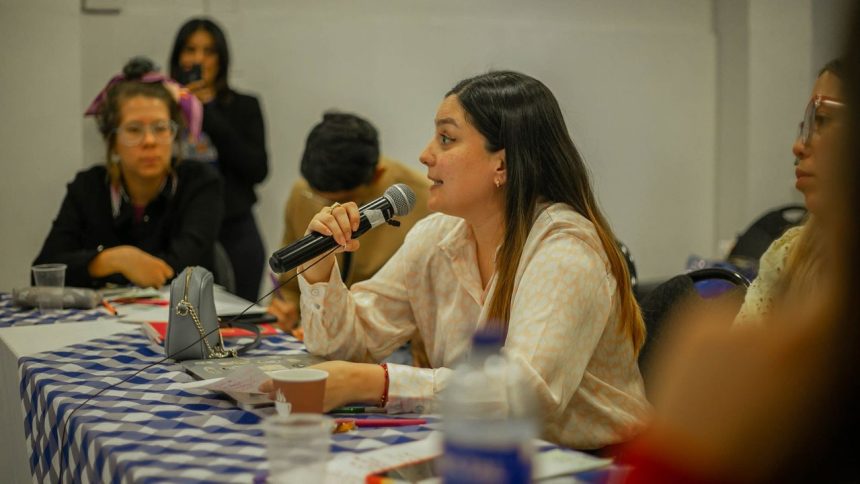 Mulher participa numa discussão de grupo, segurando um microfone junto a uma mesa.