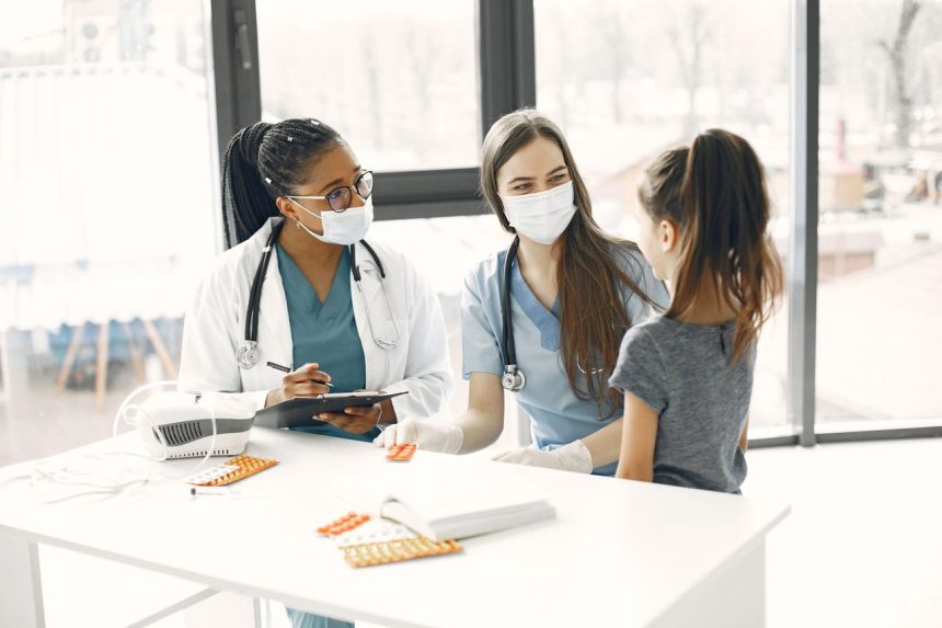 Duas médicas de máscara acompanham uma criança durante um atendimento numa sala hospitalar.