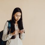 Adolescente com mochila usa um smartphone num ambiente interior minimalista.